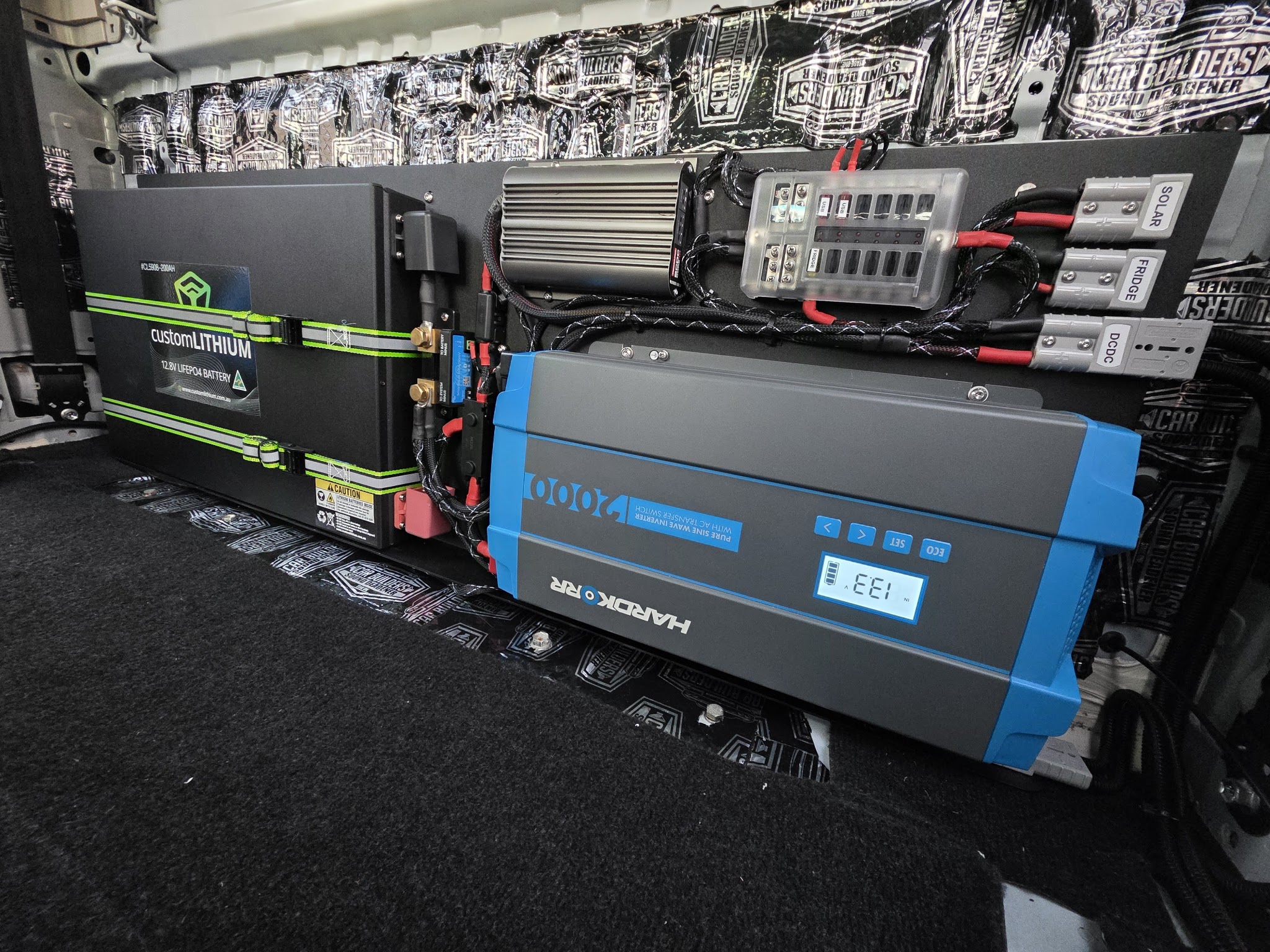 A van fitout with a Custom Lithium 12.8V battery, Hardkorr 2000W inverter charger and fuse block, on a sound-deadened wall
